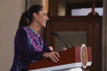 Woman in a purple outfit speaking at a podium with a seal emblem visible on the front, microphone in front of her.