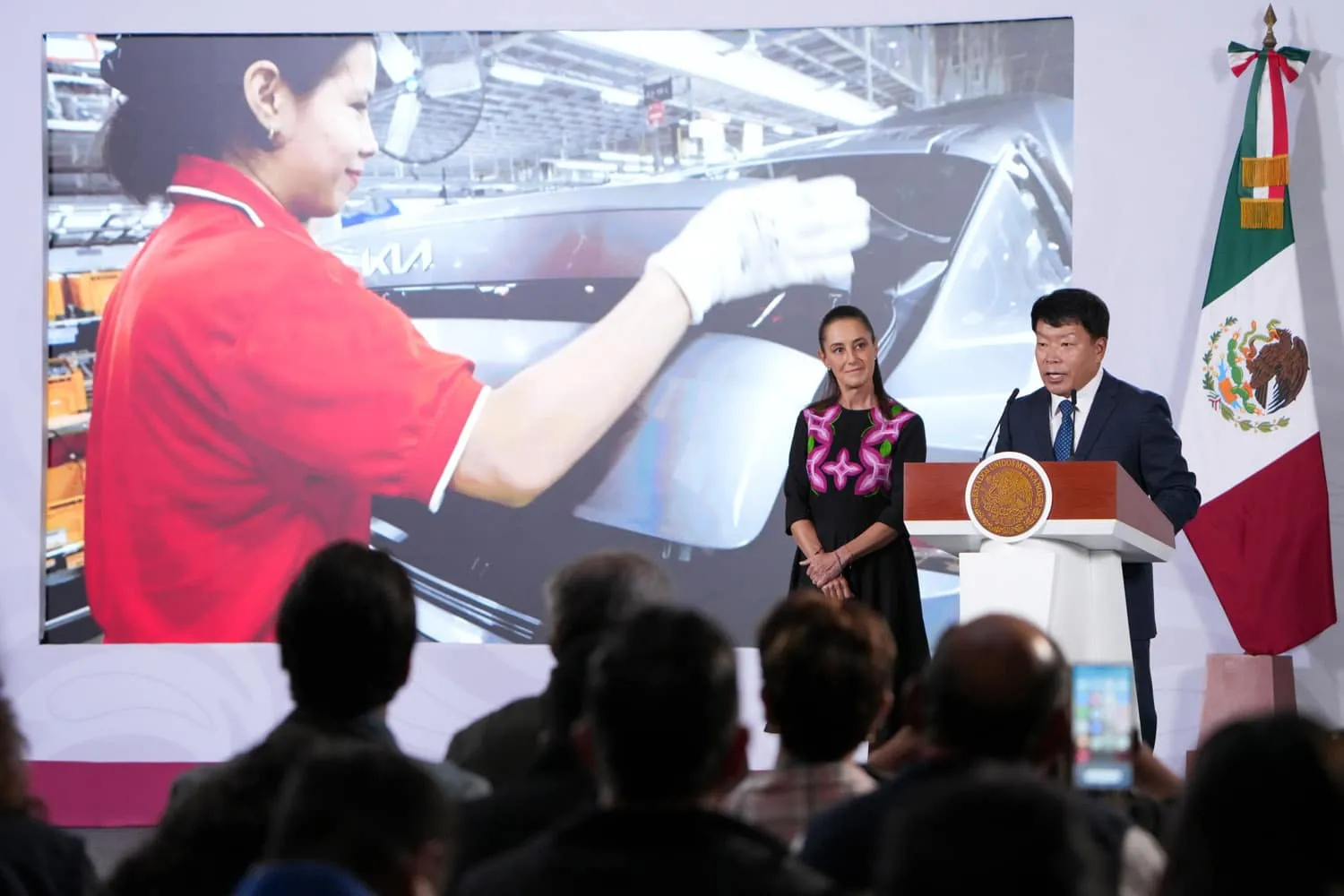 Speaker at a podium during a formal event, with a Mexican flag beside him and a woman standing nearby as a large screen shows a factory worker in a red shirt in the background.