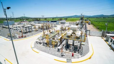 Industrial gas/processing facility with numerous yellow-handrail platforms, white pipes, and equipment set on a paved yard under a bright blue sky.