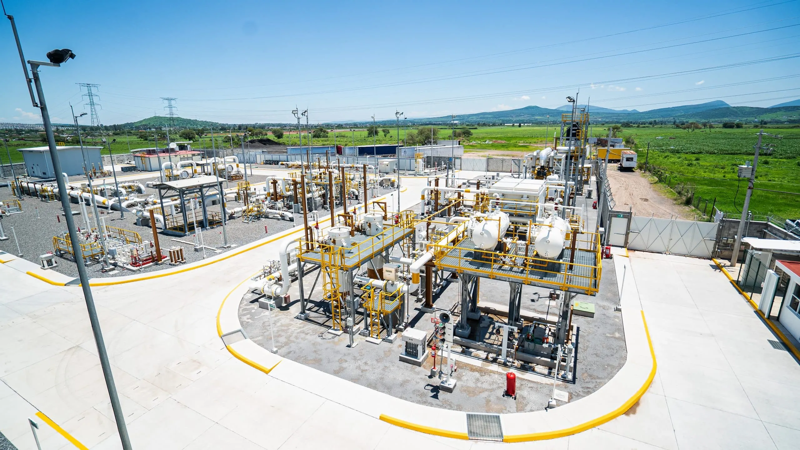 Industrial gas/processing facility with numerous yellow-handrail platforms, white pipes, and equipment set on a paved yard under a bright blue sky.