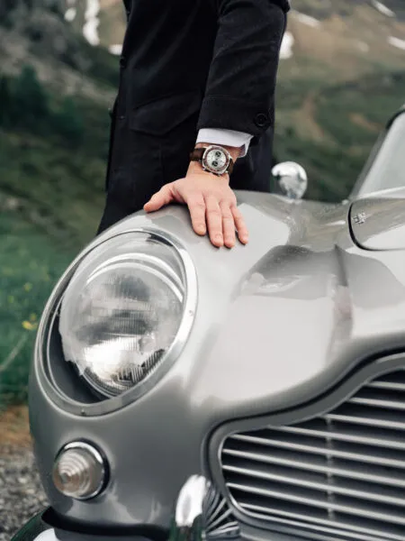 Person in a dark coat rests their hand on the hood of a vintage silver car, showcasing a brown leather–strap wristwatch on their wrist.