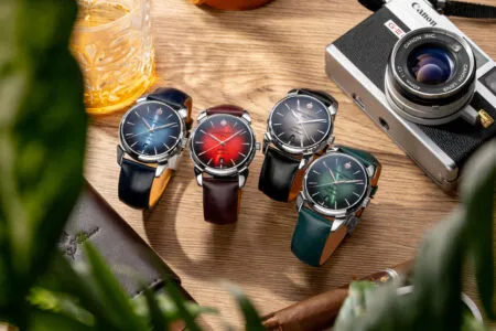 Row of colorful wristwatches with leather straps on a wooden table beside a Canon camera; foreground plants blur the edges.