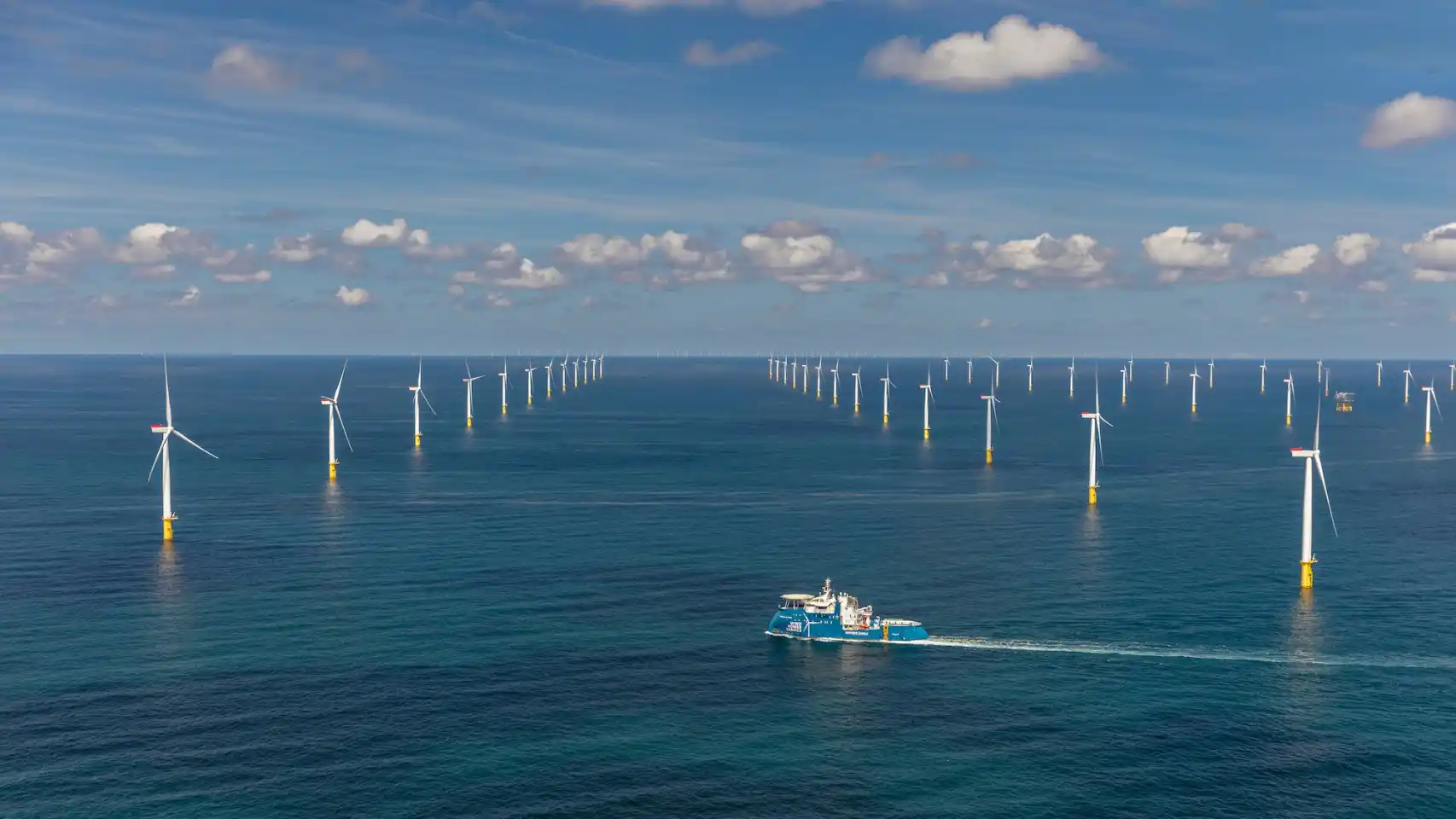 Offshore wind farm with rows of wind turbines in the blue sea and a blue support vessel nearby.