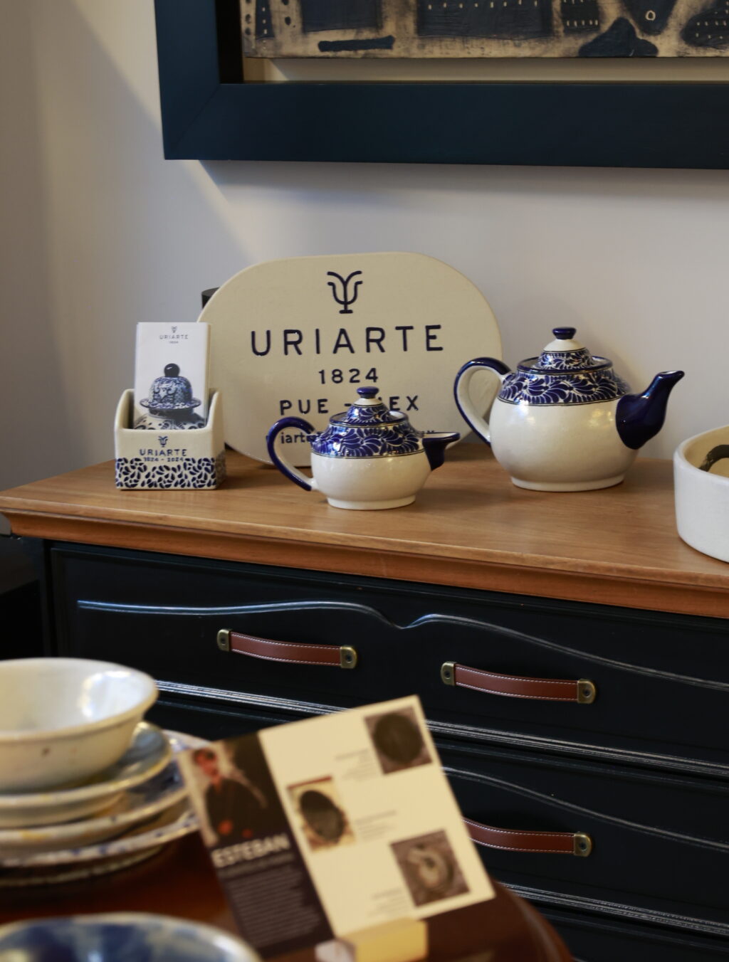 _A0A5729 - Petróleo y Energía Blue-and-white ceramic teapots and a URIARTE display sign on a wooden dresser with brochures nearby
