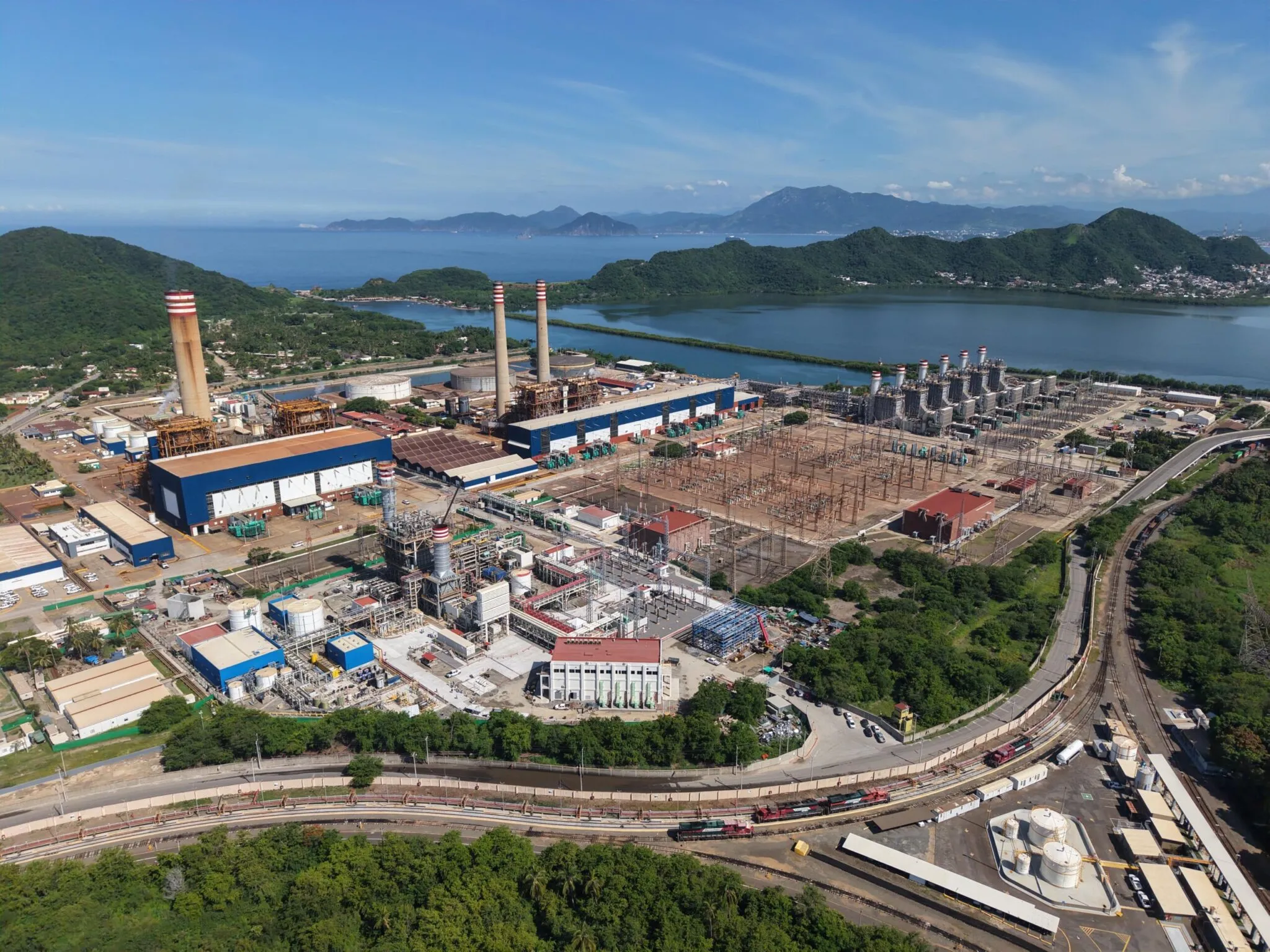 Industrial plant with tall chimneys by a blue bay, green hills in the background and distant mountains under a clear sky, daytime.