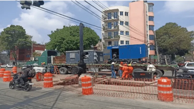 Construction crew in orange suits operates a large drilling rig on a flatbed truck at a city intersection, surrounded by orange traffic barriers and cones while a motorcyclist passes by.