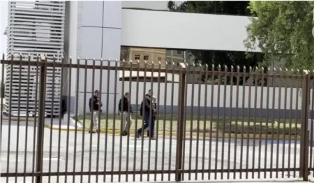Four pedestrians with backpacks walking along a sidewalk behind a tall brown metal fence near a white modern building.