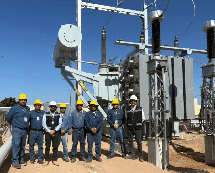 Group of utility workers in hard hats and safety vests standing in front of a high-voltage electrical substation with transformers and equipment behind them.
