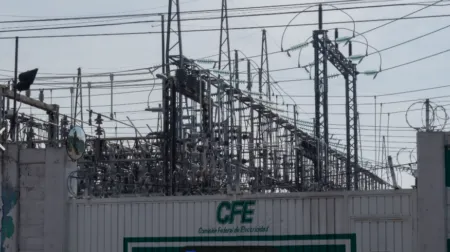 High-voltage electrical substation with transformers and a tangle of power lines behind a fence marked CFE (Comisión Federal de Electricidad) and topped with barbed wire.