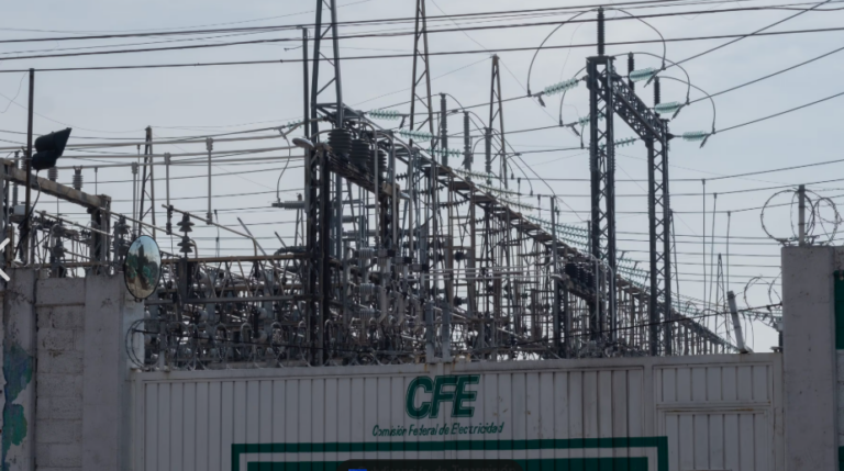 High-voltage electrical substation with transformers and a tangle of power lines behind a fence marked CFE (Comisión Federal de Electricidad) and topped with barbed wire.