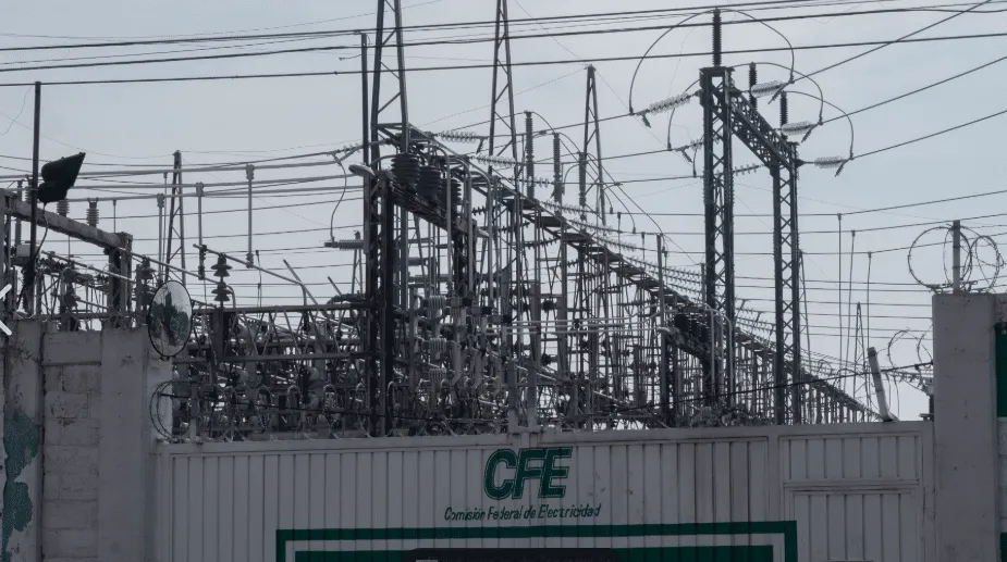 High-voltage electrical substation with transformers and a tangle of power lines behind a fence marked CFE (Comisión Federal de Electricidad) and topped with barbed wire.