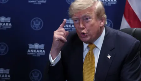 Politician in a dark suit and yellow tie, raising a finger while speaking at a press event with an Ankara backdrop behind him.