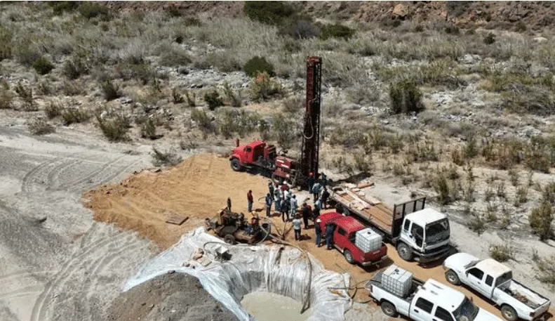 Aerial view of a desert drilling site with a tall drilling rig, red trucks, and workers around equipment.