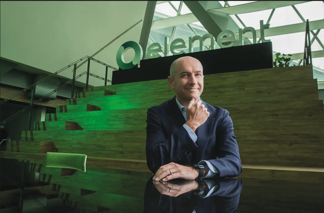 Smiling man in a navy suit sits at a glossy black table in a modern lobby with a green-lit wooden wall and a large 'element' sign behind him.