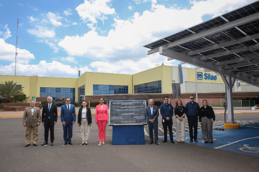 Ten professionals stand in a parking lot in front of a yellow building with a blue sign and solar canopy, posing beside a large plaque of text.