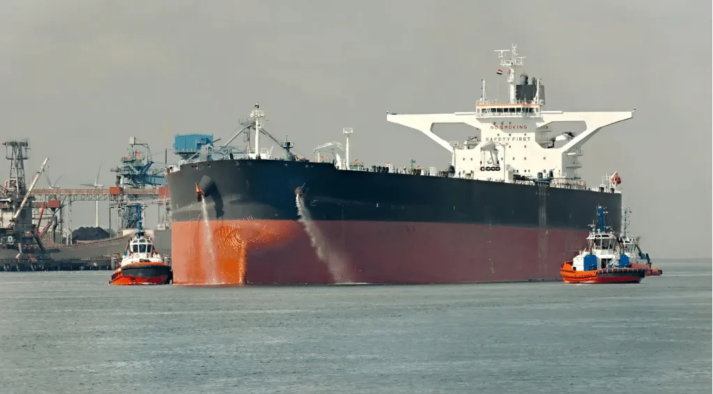 Massive bulk carrier being guided by two orange tugboats in a calm harbor.