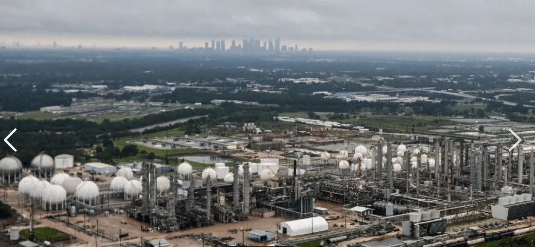 Industrial refinery site with white spherical storage tanks and tall processing towers, distant city skyline under cloudy sky.