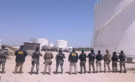 Line of armed security personnel in tactical gear standing with backs to the camera at an oil storage facility.