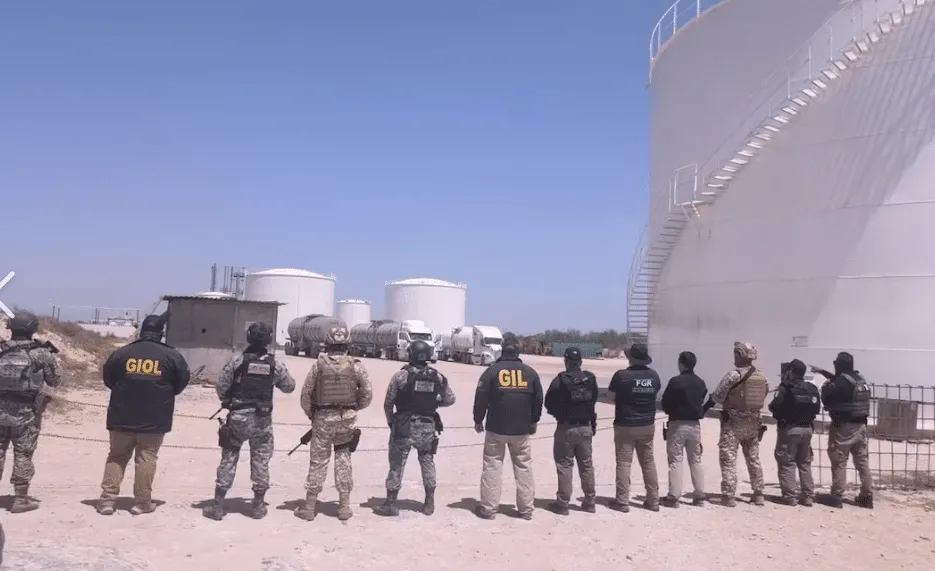 Line of armed security personnel in tactical gear standing with backs to the camera at an oil storage facility.