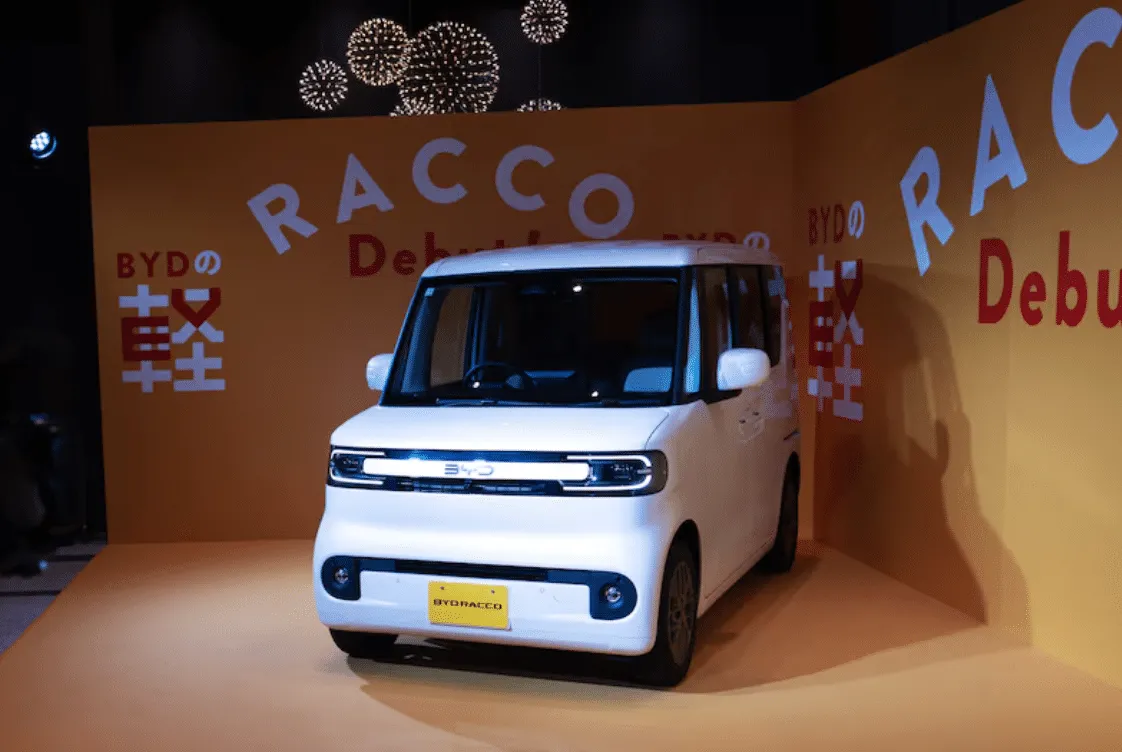 White compact van on a yellow exhibition pedestal with bold display graphics and BYD branding in the background (front view).