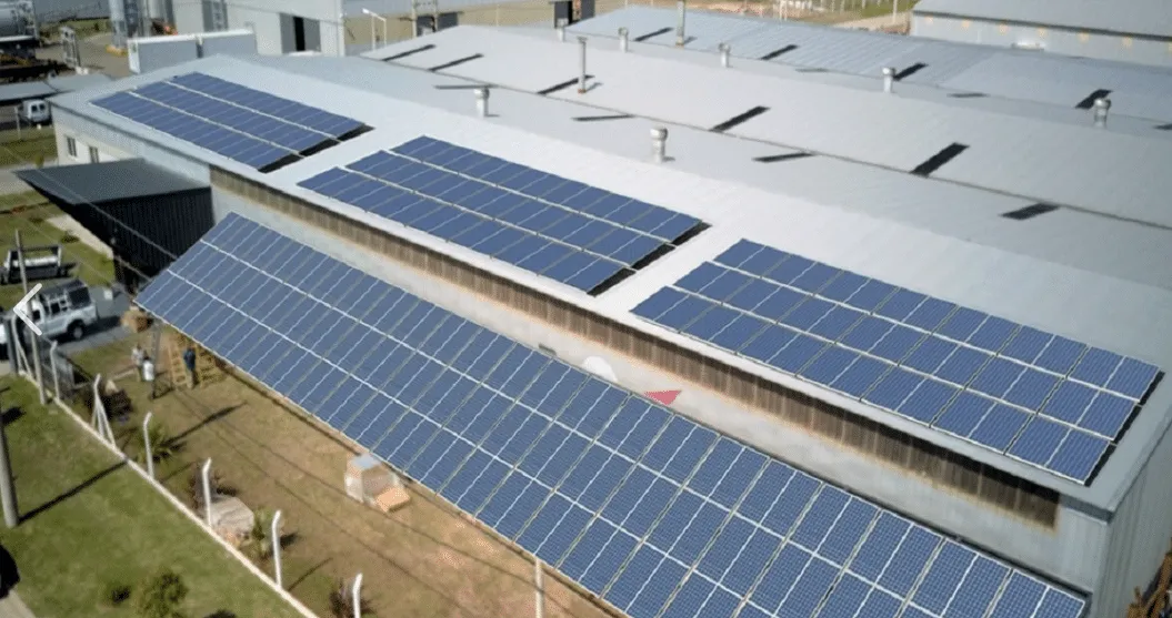 Aerial view of solar panels installed on the roof of an industrial warehouse, arranged in long rows on a metal roof.