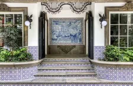 Entrance with blue tiled mural framed by iron gates and stone molding, flanked by plants and bushes.