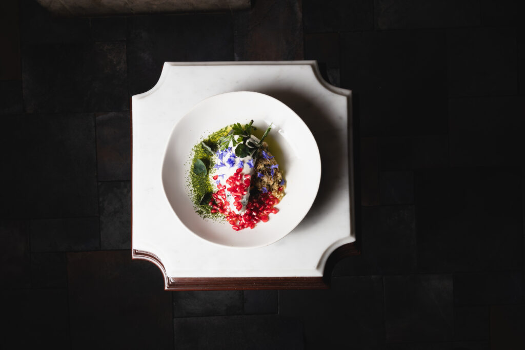 ChileNogada_LaFridaCabo_1 - Petróleo y Energía Top-down view of a white plate on a small white stand, garnished with red seeds, green powder, and purple edible flowers against a dark floor.