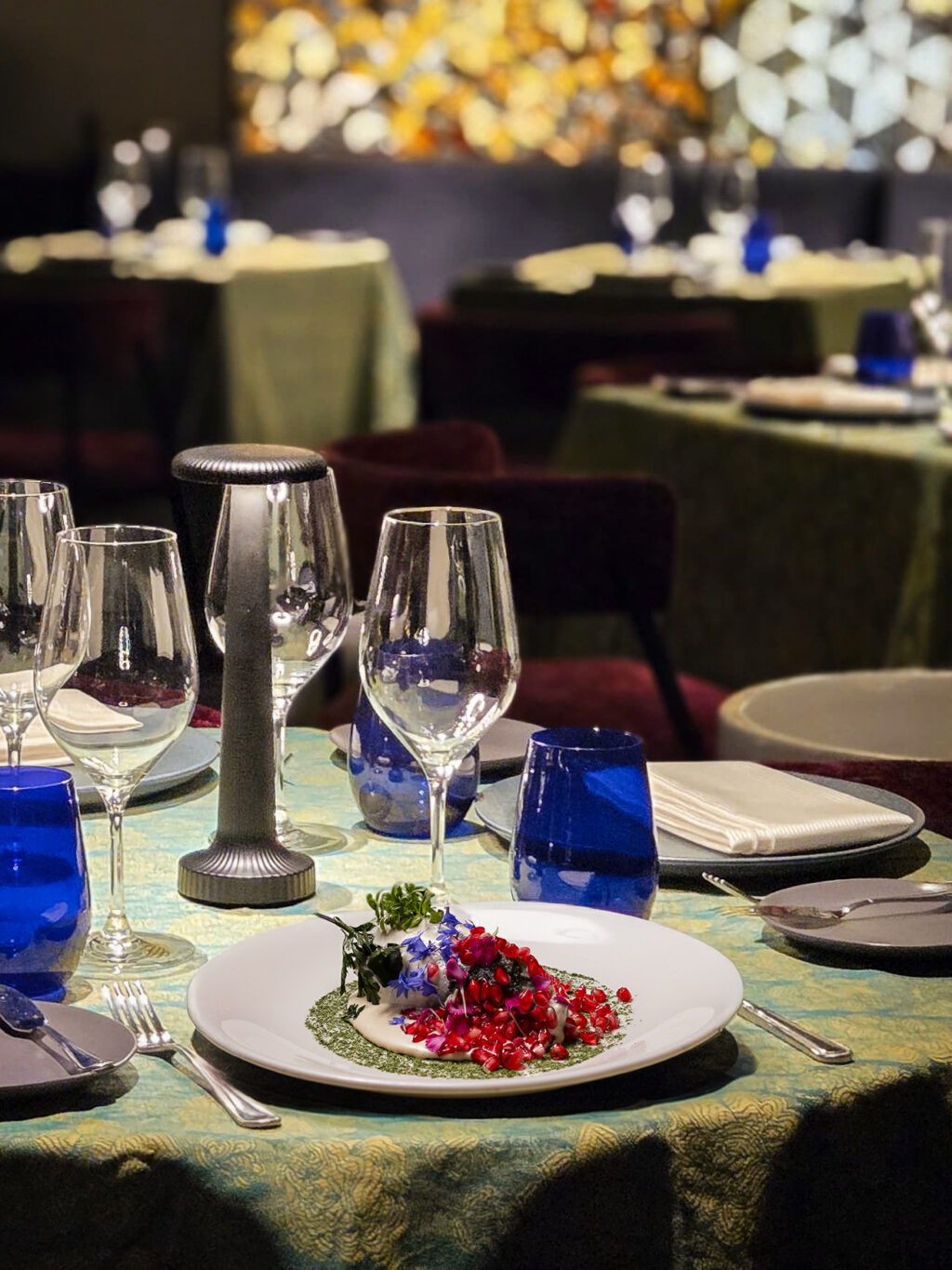 ChileNogada_LaFridaSMA_1 - Petróleo y Energía Formal dining table set with blue glassware, wine glasses, and a plated dessert garnished with pomegranate seeds and herbs.