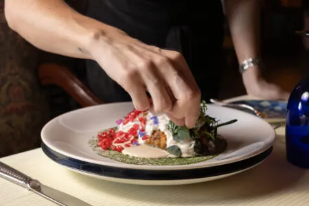 Hands garnish a plated dessert with red seeds and green herbs on a white plate at table level.
