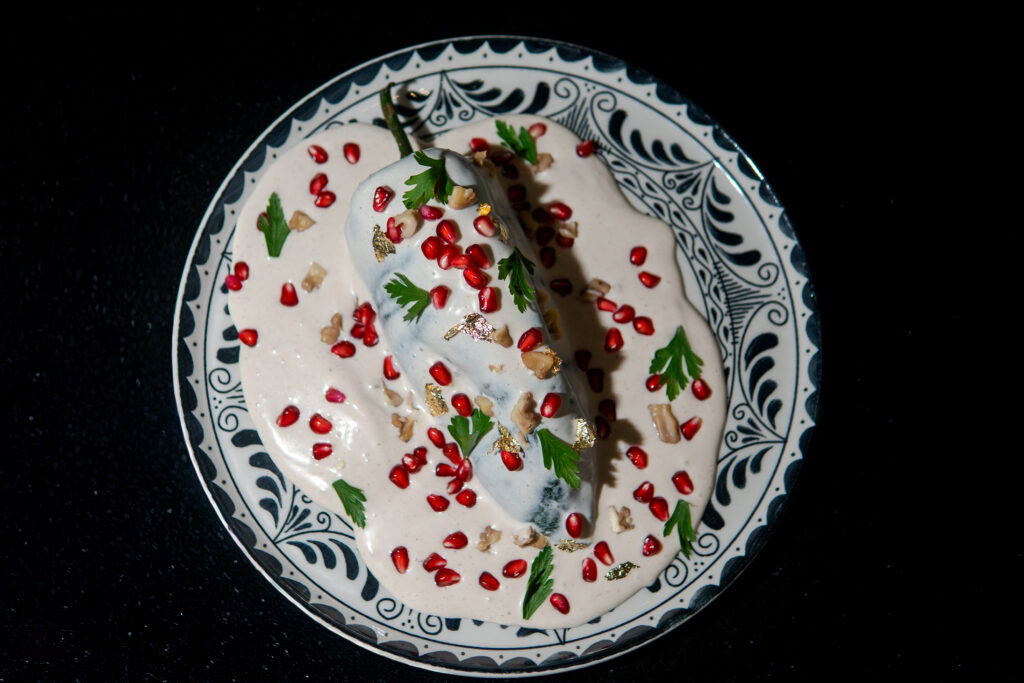 ChileNogada_Melville_4 - Petróleo y Energía Creamy white dessert on a decorative blue-and-white plate, topped with pomegranate seeds, parsley, chopped nuts, and gold leaf on a dark background.