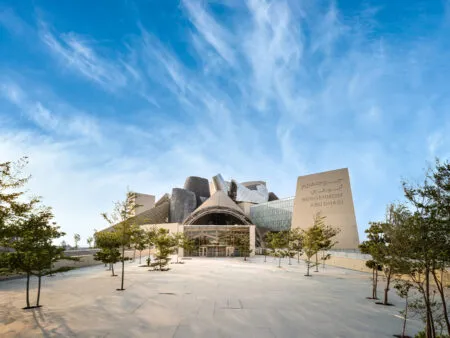Exterior view of the Guggenheim Abu Dhabi museum with sculptural metal panels and a wide, paved forecourt under a blue sky, surrounded by trees