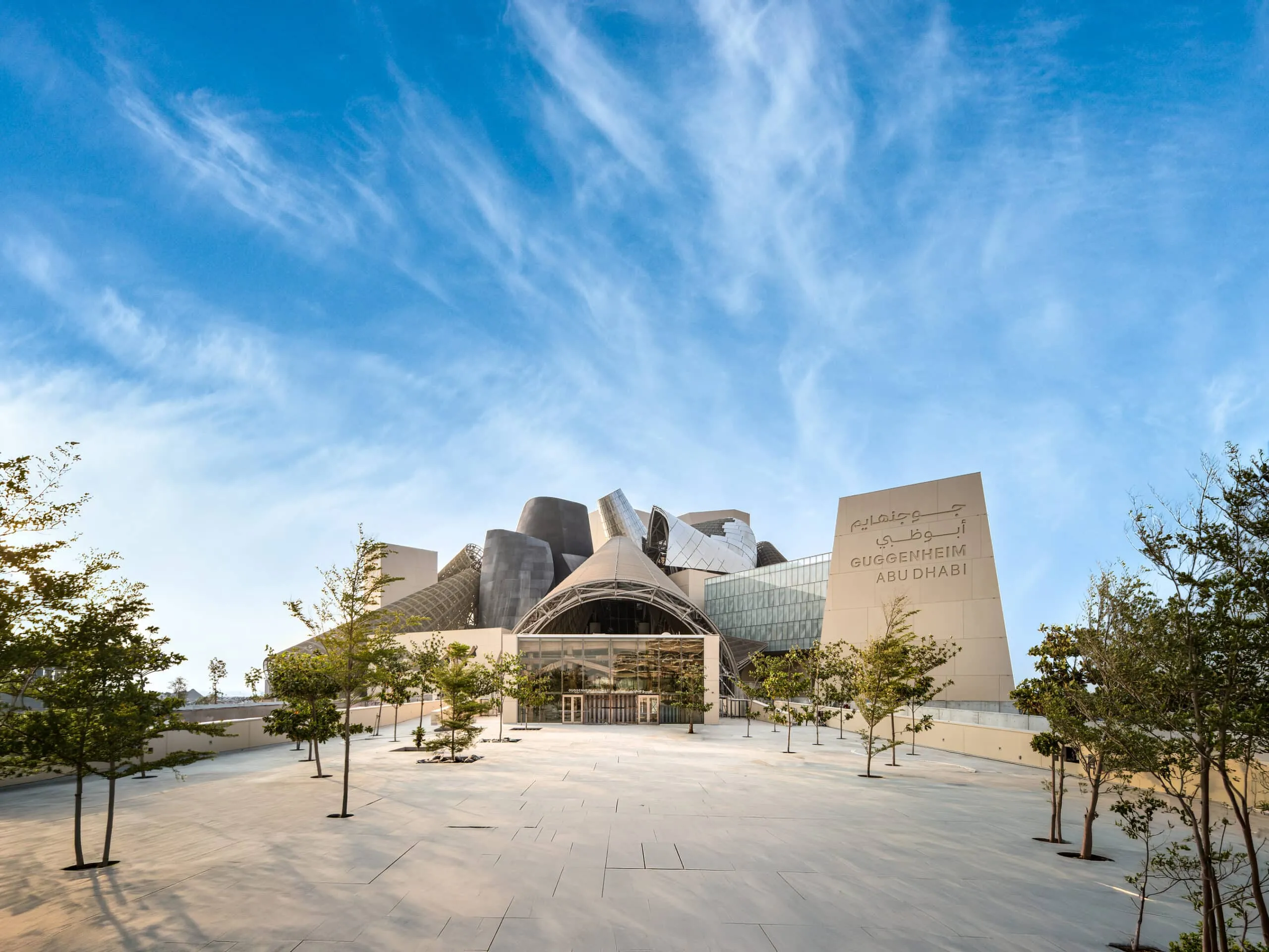 Exterior view of the Guggenheim Abu Dhabi museum with sculptural metal panels and a wide, paved forecourt under a blue sky, surrounded by trees