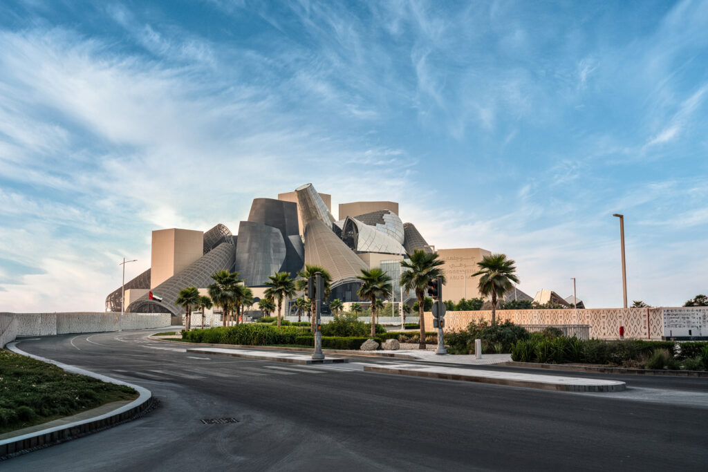 DCT-Abu-Dhabi-4-Guggenheim-Abu-Dhabi - Petróleo y Energía Exterior view of the Guggenheim Abu Dhabi with sculptural, silver metal forms against a blue sky and palm-lined foreground road.