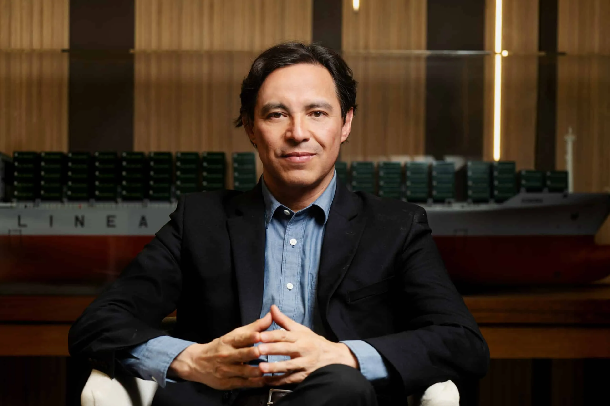 Professional portrait of a man in a dark blazer and blue shirt, seated with hands folded, in a wood-paneled conference room.