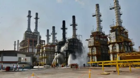Industrial refinery with tall distillation columns; firefighters spray water at center of the scene.