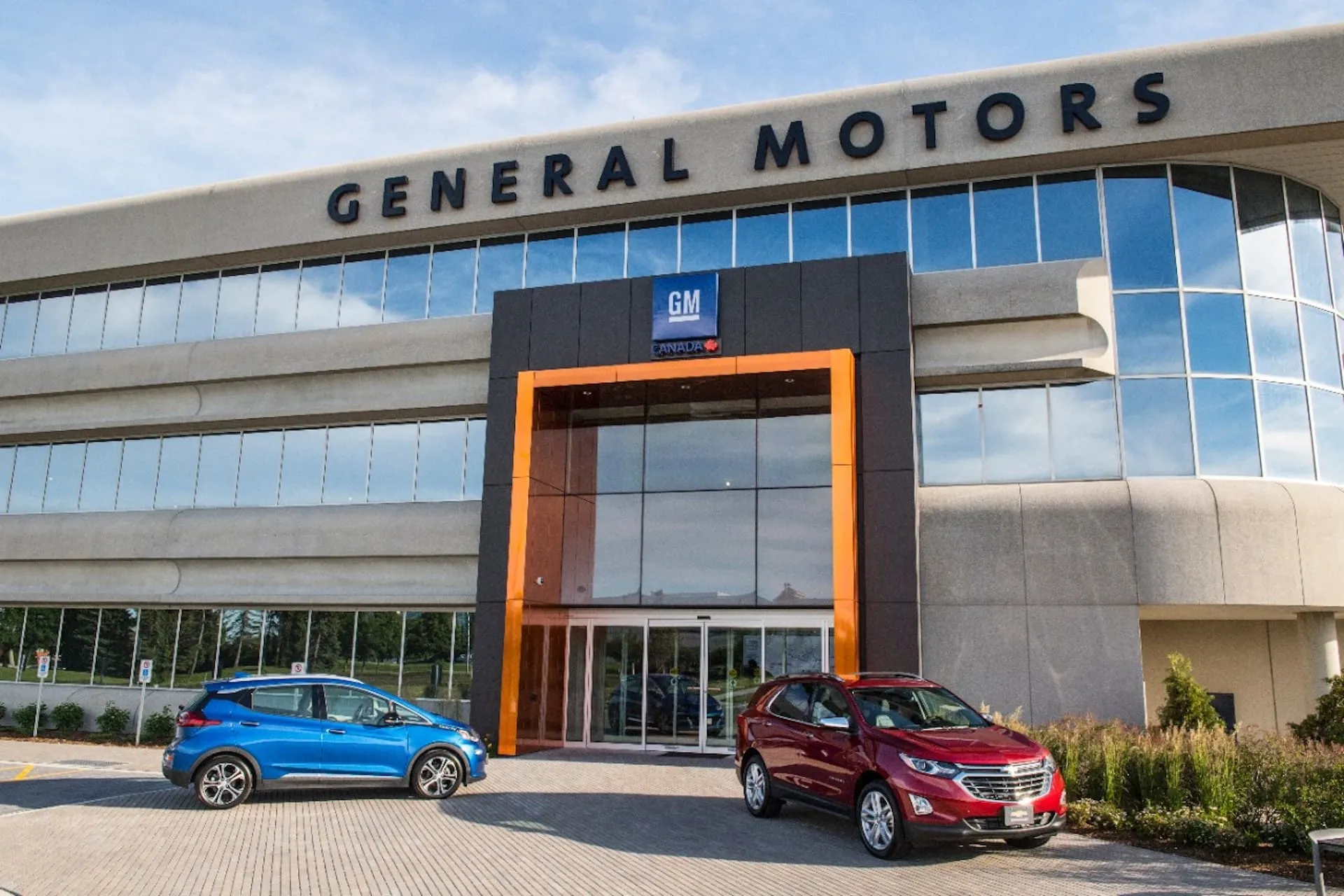 Front view of a General Motors office building with glass walls, an orange-framed entrance, and blue and red cars parked outside.