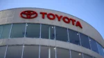 Toyota dealership building with red logo and 'TOYOTA' signage on curved glass facade against a blue sky.