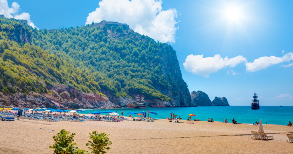 Guide-to-Cleopatra-Beach-in-Alanya - Petróleo y Energía Sunlit sandy beach with blue umbrellas, turquoise sea, and green cliffs in the background.