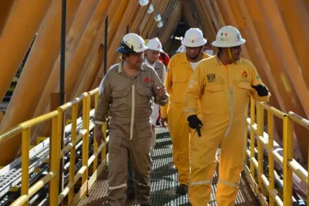 Group of construction workers in hard hats and jumpsuits walk along a yellow steel catwalk between large orange support beams on a sunny day.