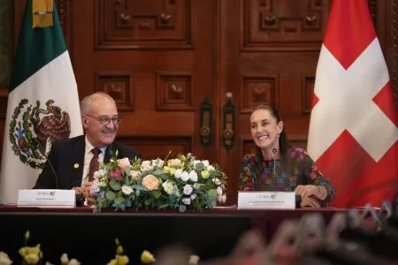 Two officials sit at a ceremony table with a large flower arrangement, Mexico on the left and Switzerland on the right flag backdrop.