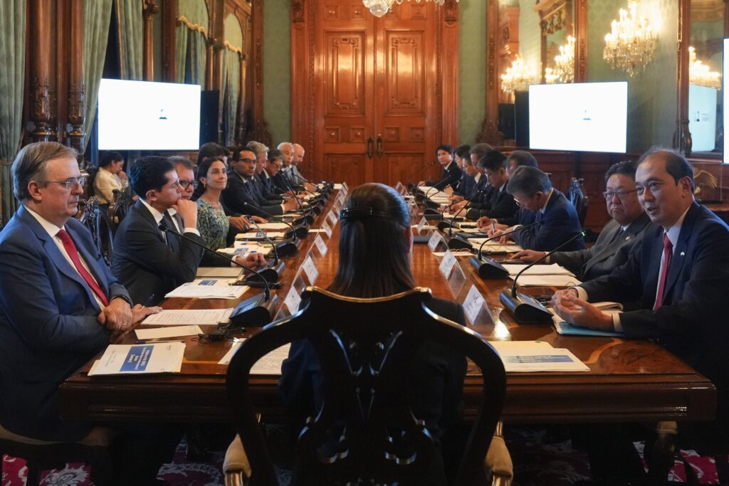 HNJ4DDGW8AAhvO2 - Petróleo y Energía Long conference table with delegates in formal attire seated on both sides, microphones and documents before them in a grand wood-paneled room.