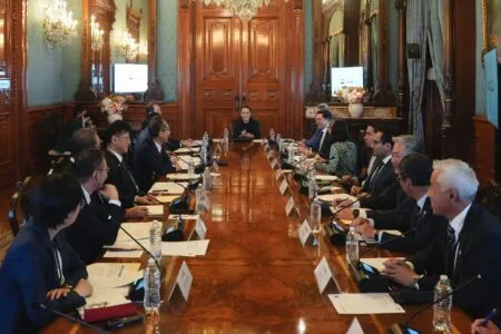 Formal boardroom meeting with a woman speaking at the head of a long wooden table in an ornate room with chandeliers and wood paneling