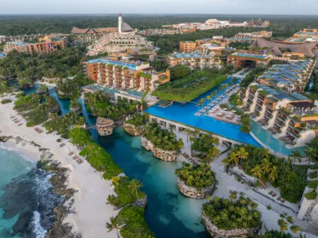 Aerial view of a tropical resort with turquoise lagoons, bridges, and beachfront towers surrounded by palm trees and greenery.