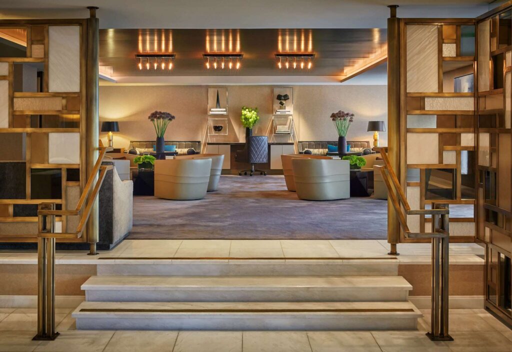 LErmitage Beverly Hills Spa - Petróleo y Energía Symmetrical hotel lobby with beige sofas, round chairs, and warm gold lighting seen through decorative wooden screens.