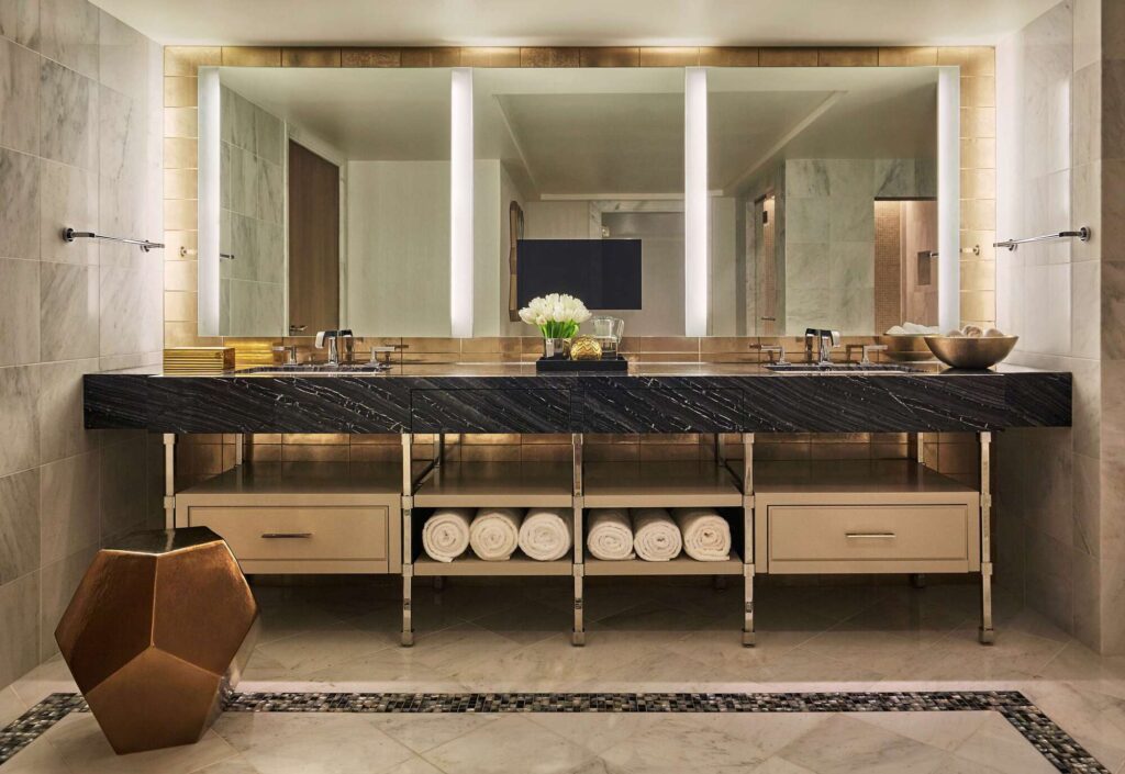 LErmitage Beverly Hills Spa 2 - Petróleo y Energía Luxurious bathroom vanity with black marble countertop, double sinks, and bright mirrors. A small vase of white flowers sits in the center.
