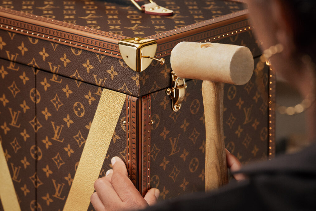 LOUIS_VUITTON_FIFA_WORLD_CUP_2026_TROPHY_TRUNK_SAVOIR_FAIRE_VISUALS15M - Petróleo y Energía Close-up of a brown Louis Vuitton trunk with a gold lock and a wooden mallet resting on the corner.