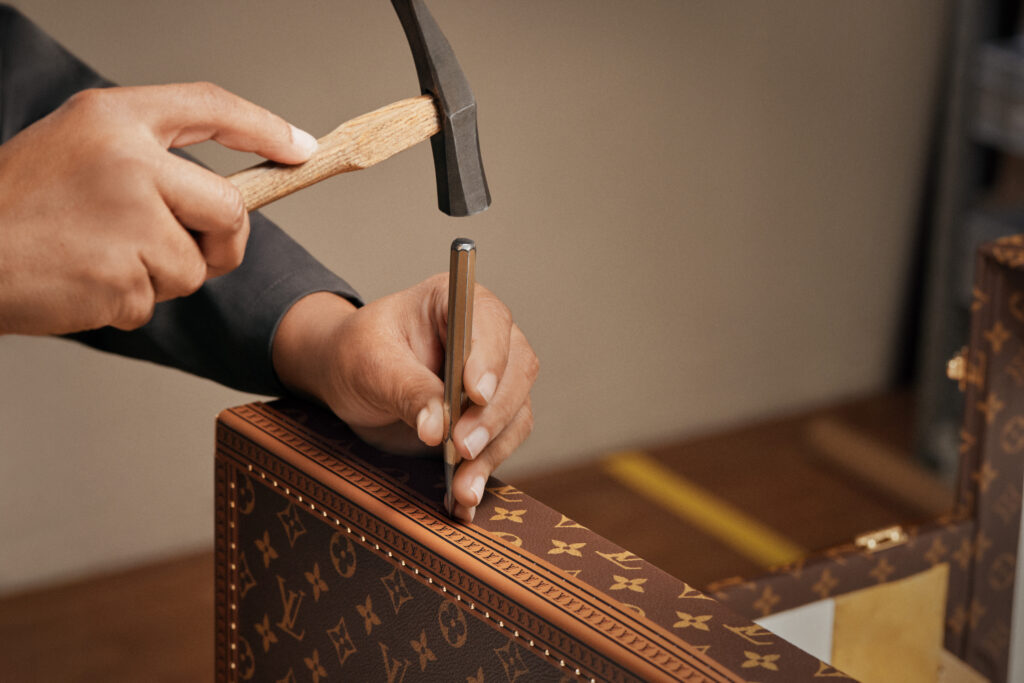 LOUIS_VUITTON_FIFA_WORLD_CUP_2026_TROPHY_TRUNK_SAVOIR_FAIRE_VISUALS2M - Petróleo y Energía Close-up of hands using a hammer and chisel to carve along the edge of a patterned brown box surface.