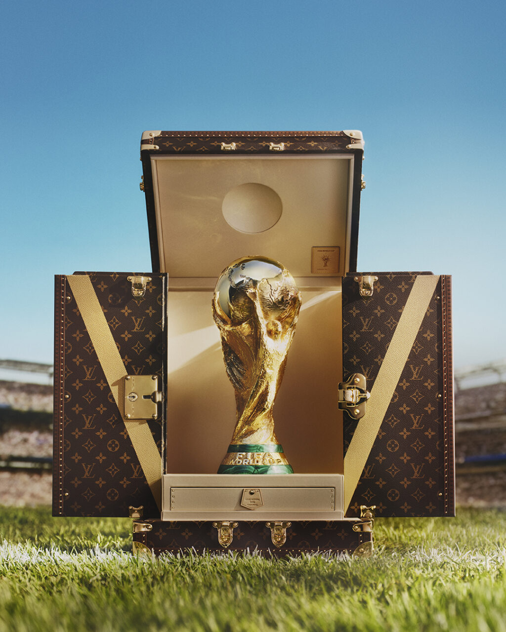 LOUIS_VUITTON_x_FIFA_WORLD_CUP_2026_TROPHY_TRUNK_C - Petróleo y Energía Golden World Cup trophy nestled inside an open luxury LV suitcase, set on grass with a stadium backdrop under a blue sky.