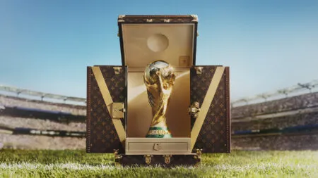 Golden World Cup trophy rising from an open Louis Vuitton suitcase on a football pitch, stadium full of spectators in the background.