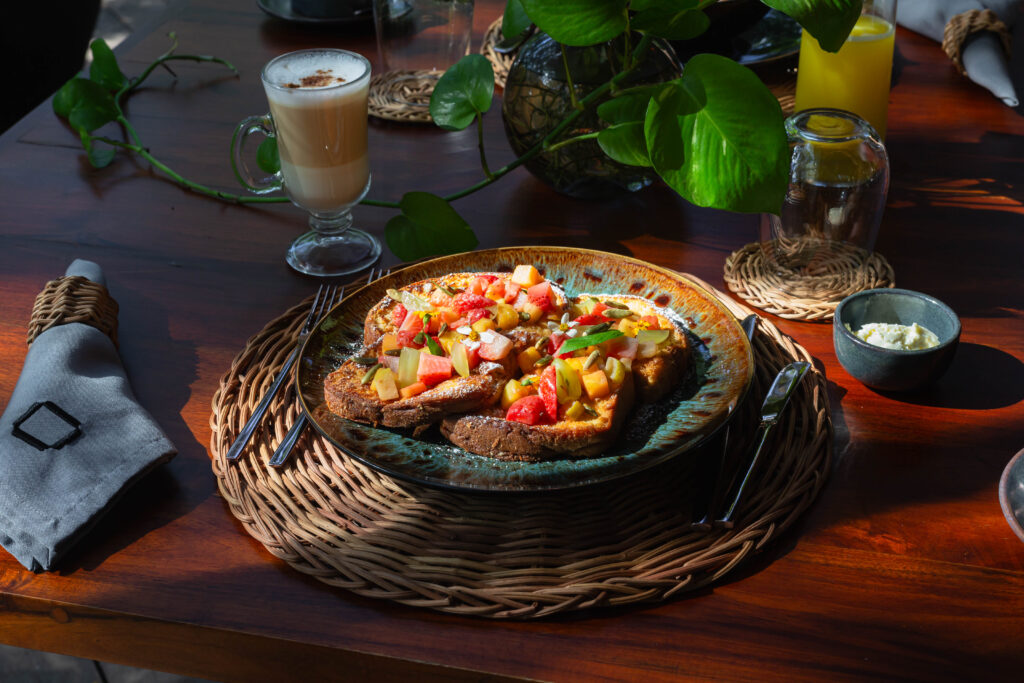 MESTIZO MISTERIO CULINARIO 3 - Petróleo y Energía Plate of fruit-topped French toast on a blue ceramic plate with a woven placemat on a wooden table; latte in a glass mug and greenery in the background.